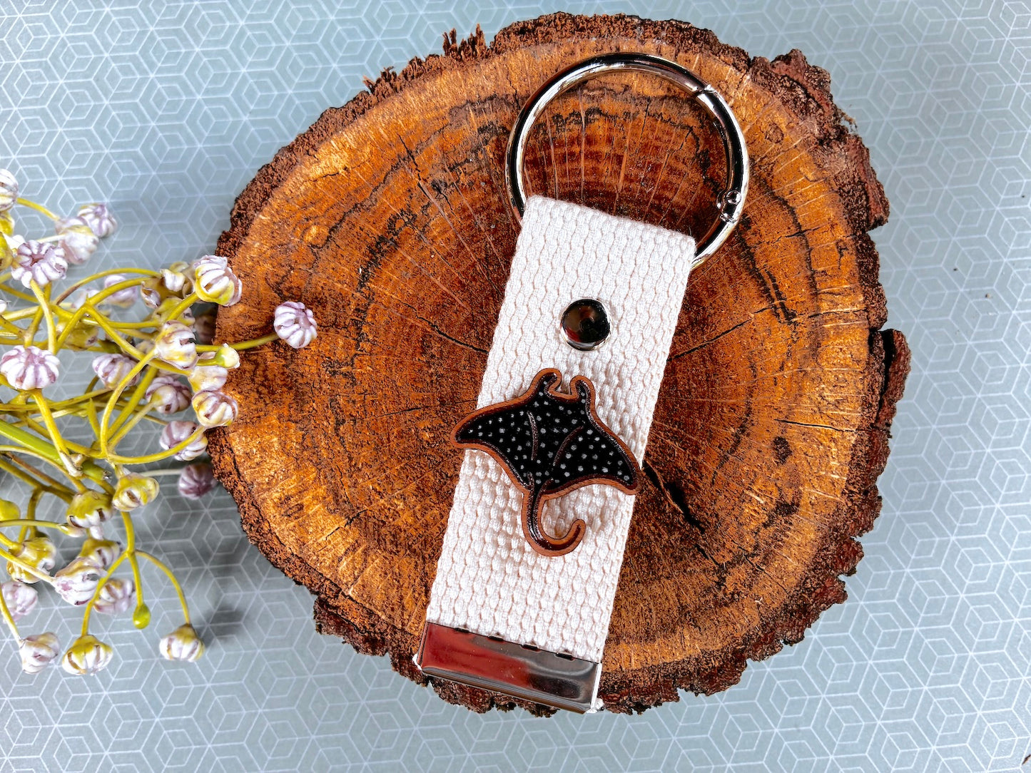 A white woven fabric keychain with a black and white manta ray charm, resting on a rustic wood slice with small white flowers on a light blue patterned background.
